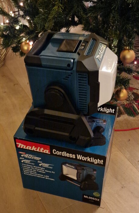 Makita sladdlös arbetslampa ML005GX, placerad på sin förpackning, med dekorerad julgran i bakgrunden.