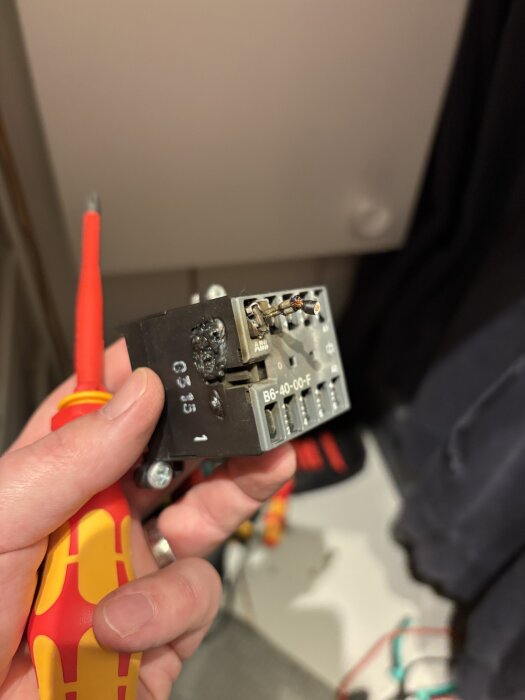 Närbild på en hand som håller en kontaktor, bredvid en skruvmejsel. Kontaktorn är delvis skadad eller bränd.