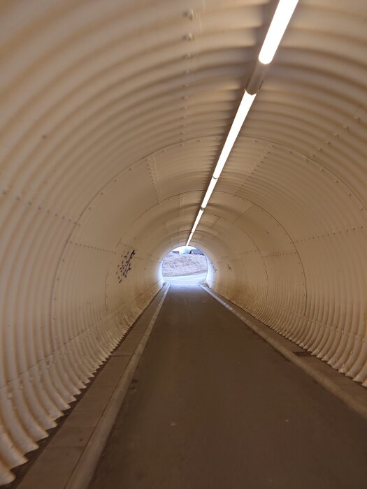 Gångtunnel med korrugerade väggar och tak, upplyst med lysrör, leder ut till ett öppet område i dagsljus.