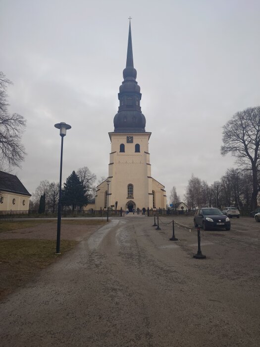 Kyrka med hög spira och klocka på en mulen dag, omgiven av träd och bilar, med människor på vägen mot ingången.