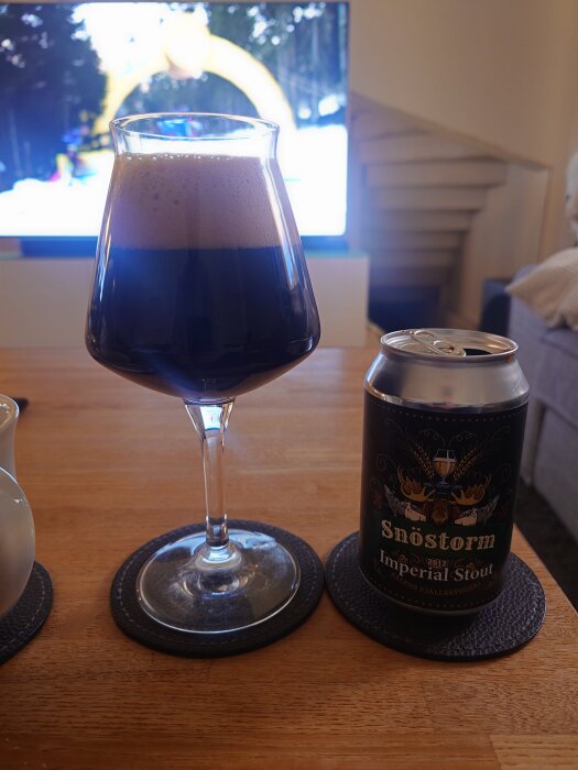 Ett glas Snöstorm Imperial Stout från Sälens bryggeri står på ett bord bredvid en öppen ölburk, med en tv i bakgrunden.