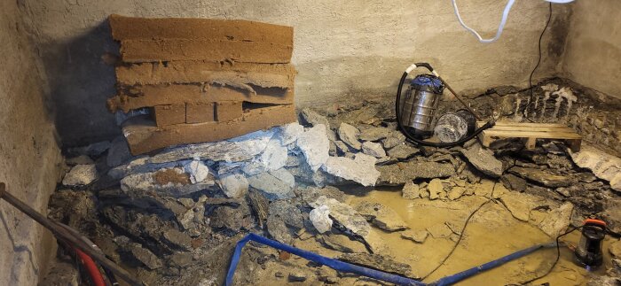 Källarprojekt med upplagda stenblock, sand, en vattendammsugare och diverse verktyg spridda på golvet och längs väggen.
