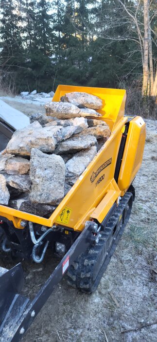 Gul minidumper lastad med stenar står på en skogstomt.
