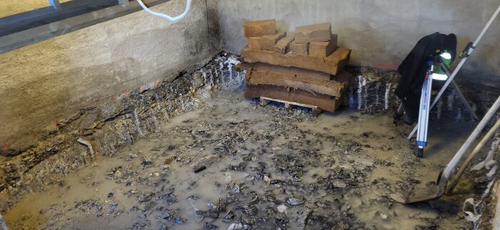 Bilden visar en källare under renovering med grus och sten spillror på golvet, staplade jordblock längs väggen, och en spade i hörnet.