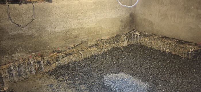 Källarhörna med cementvägg och singel på golvet, marken jämnad efter rensning av sten. Före renovering längs väggarna.