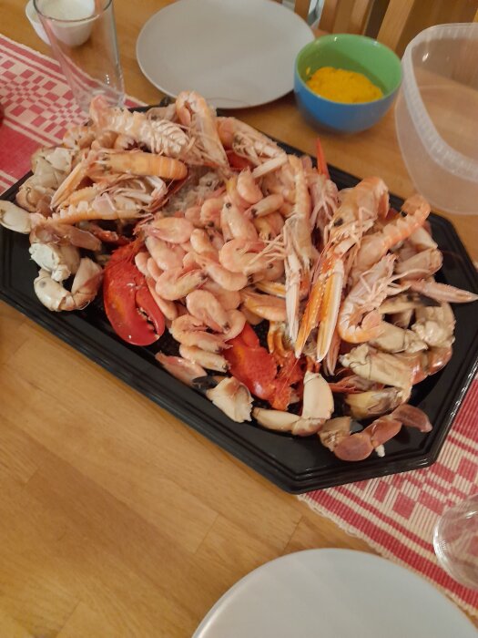 Ett fat med skaldjur som räkor, kräftor och krabbor på ett träbord, omgivna av tallrikar och en grön skål med gul sås i bakgrunden.