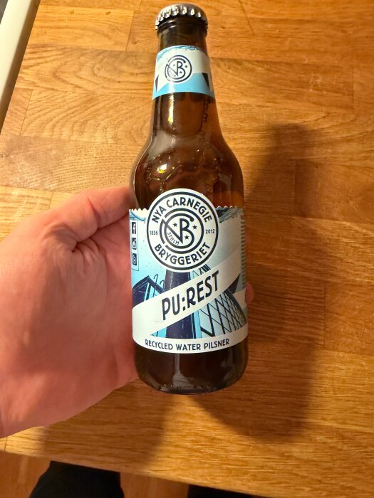 En hand håller en flaska öl från Nya Carnegie Bryggeriet, med etiketten "Pu:rest Recycled Water Pilsner" på en träbänkskiva.