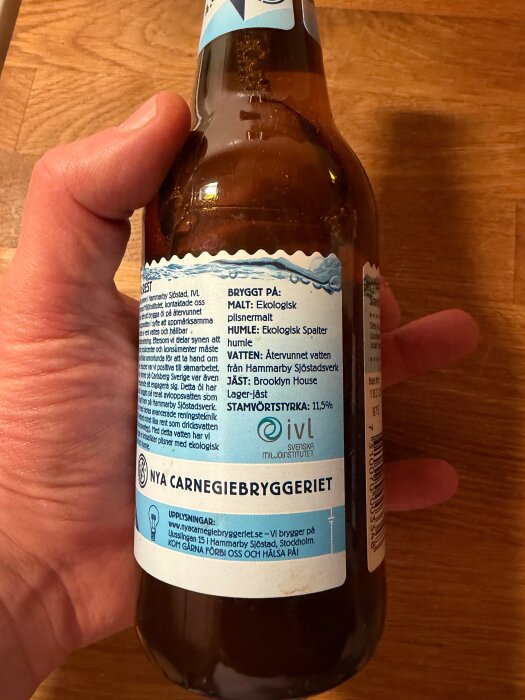 En hand håller en ölflaska från Nya Carnegiebryggeriet med etikett om ingredienser och bryggeriinformation.