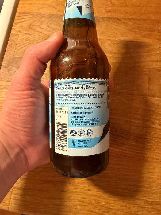 En hand håller i en ölflaska med etikett som visar alkoholhalt 4,8%, volym 33 cl och information om tillverkare och innehåll på en träyta i bakgrunden.