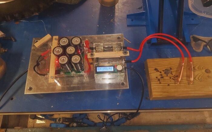 Batterisvets med batterier och digital display på en arbetsbänk, ansluten med röda kablar och tråd på träplatta.