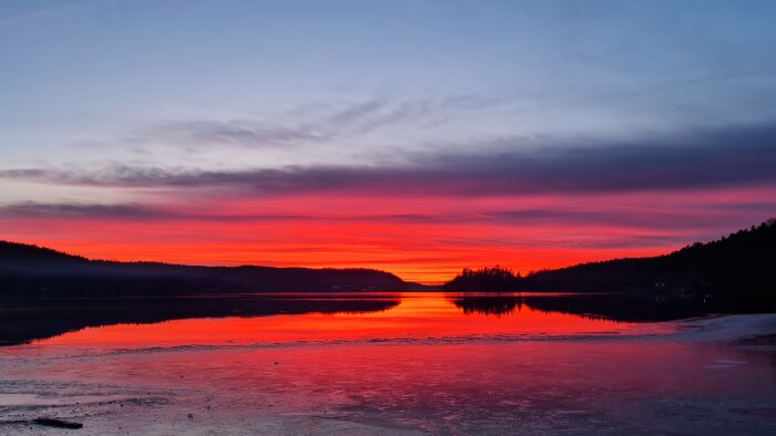 Dramatisk solnedgång med glödande röd och orange himmel reflekterar i en lugn sjö, omgiven av mörka silhuetter av träd och berg.
