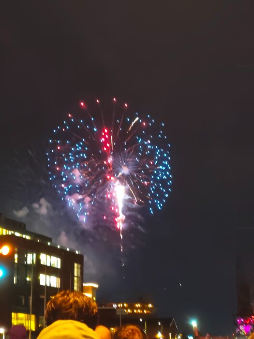 Fyrverkerier exploderar på natthimlen över en stad, med folk som tittar på från gatan nedanför.