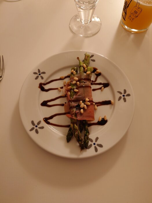Sparris inlindad i prosciutto med pinjenötter och balsamvinäger på en vit tallrik, serverad med en dryck i bakgrunden.