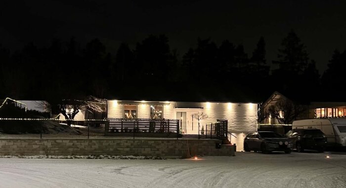 En villa upplyst av fasadbelysning och dekorativa ljusslingor på natten, omgiven av snötäckt mark och parkerade bilar.