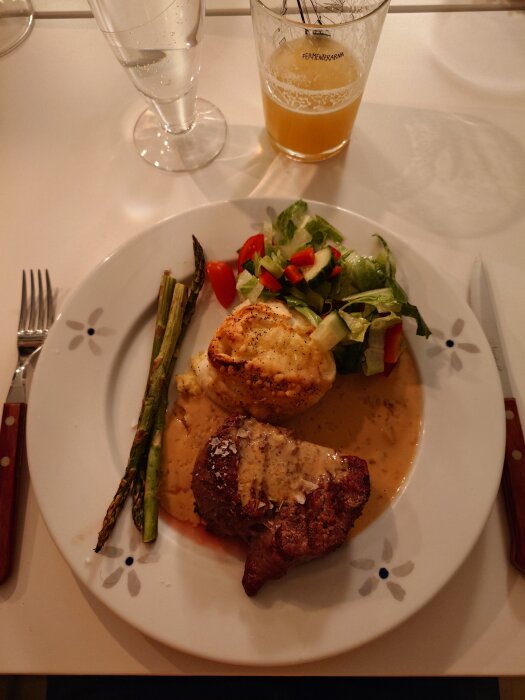 Tallrik med köttbit, potatisgratäng, sparris och sallad, serverad med sås. Ett glas öl och vatten står bredvid på bordet.