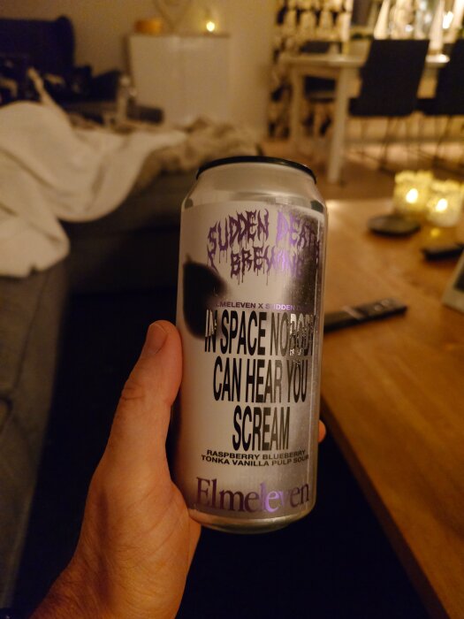 En hand håller en burk öl med etiketten "In Space No One Can Hear You Scream" från Elmeleven x Sudden Death Brewing i ett vardagsrum.