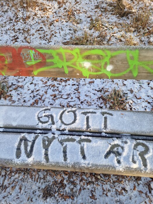 Snöig bänk med "Gott Nytt År" skrivet på den, omgiven av löv och mark täckt av snö. Graffiti i grönt och orange syns på bänken ovanför.