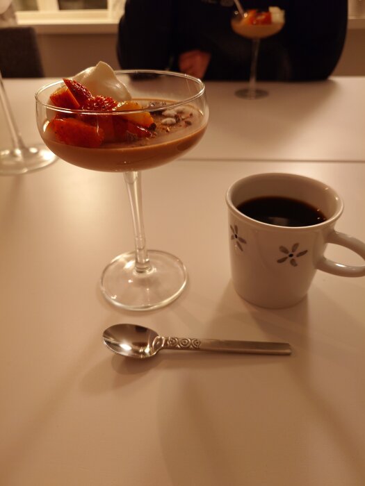 Daim pannacotta toppad med jordgubbar och grädde serverad i glas, bredvid en kopp kaffe och en sked.