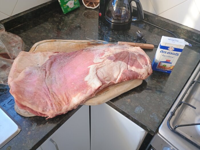 Rått lammkött med salt på en skärbräda bredvid en grillkniv och ett paket grovsalt på en köksbänk.