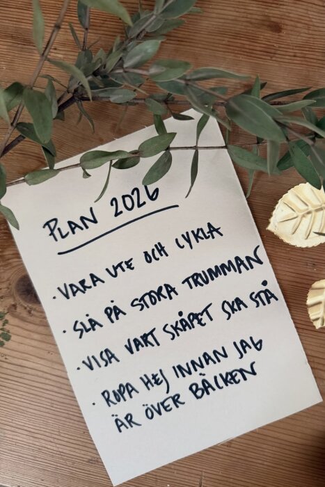 Vit papper med texten "Plan 2026" och fyra punkter på svenska, omgiven av gröna blad och gul dekoration på träbord.