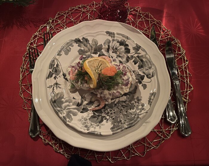 Skagenbakelse med citron, dill och rom på blommönstrad tallrik, serverad på röd duk med bestick och glasunderlägg.