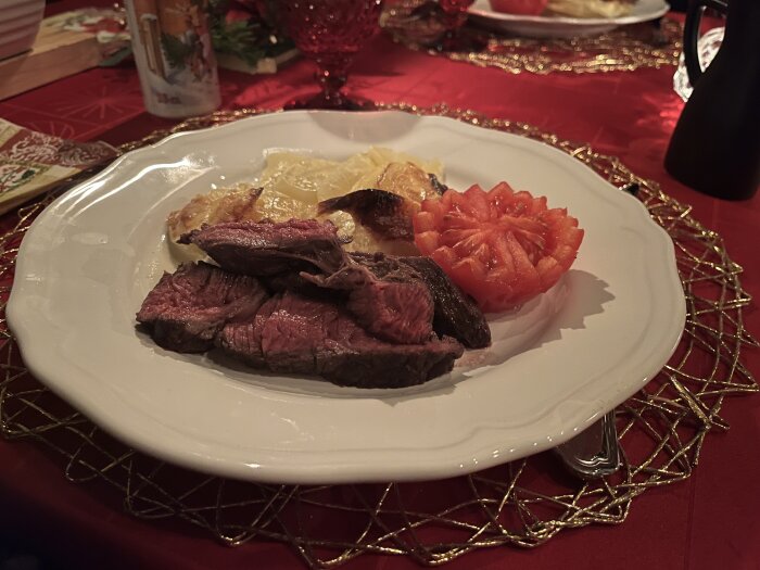 Entrecôte med potatisgratäng och skivad tomat på en vit tallrik, serverad på en röd duk.