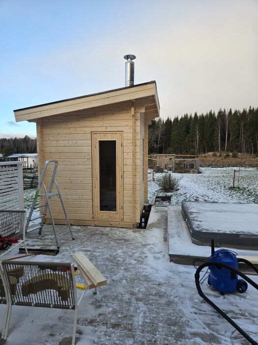 Bastu byggd på altandäck med skorsten, omgiven av snö. Stege och verktyg syns bredvid. Skog i bakgrunden.