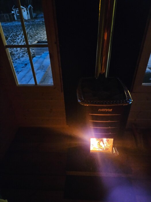 Bastukamin i bruk med eld inuti, sedd från insidan av en bastu i trä, med delvis snötäckt altan synlig genom fönstret.