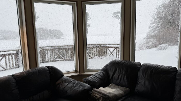 Utsikt frÄn vardagsrum över snötÀckt landskap genom stora fönster, mörka soffor i förgrunden.