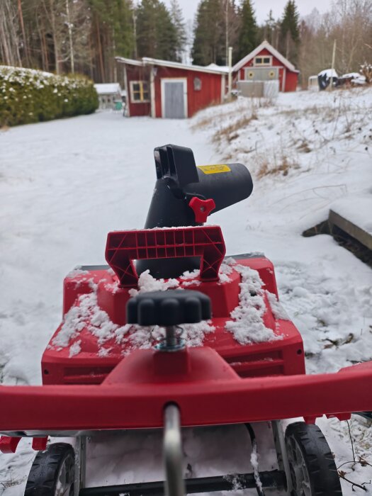 Röd snöslunga används för att röja snö på en uppfart, med röda byggnader och skog som bakgrund.