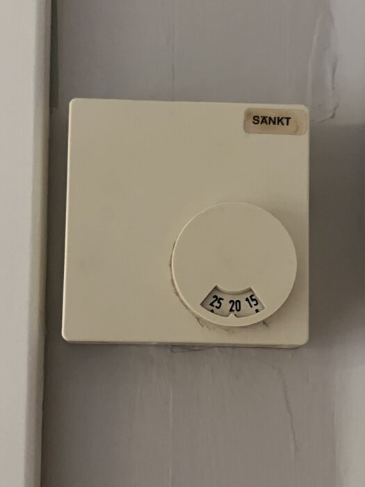 En termostat på en vägg med inställningar markerade 25, 20 och 15 grader, etikett "SÄNKT" synlig i hörnet.
