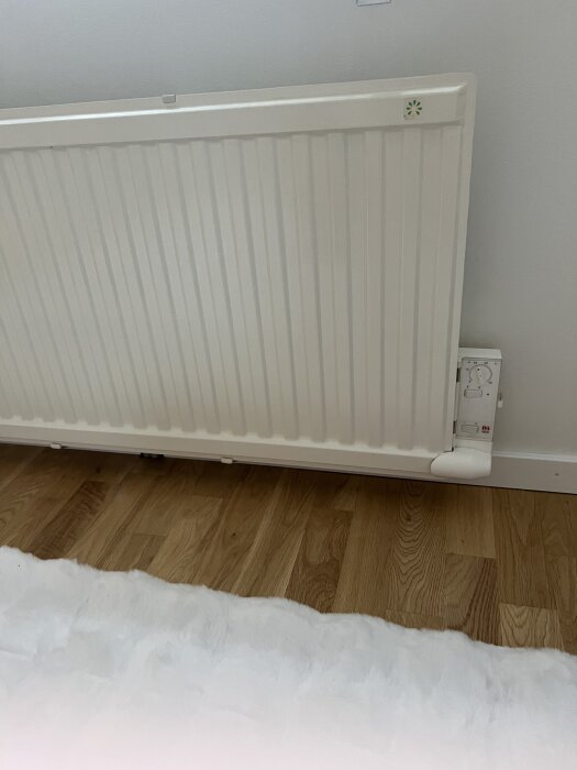 Vit elradiator mot vägg med termostat nederst installeras ovanför parkettgolv.