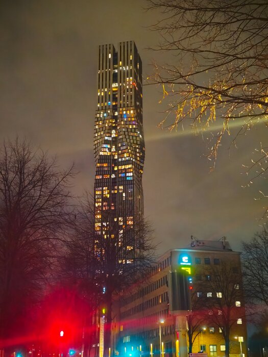 En nattbild av en upplyst skyskrapa med fönster i olika färger, omgiven av träd och en gata med trafikljus i förgrunden.