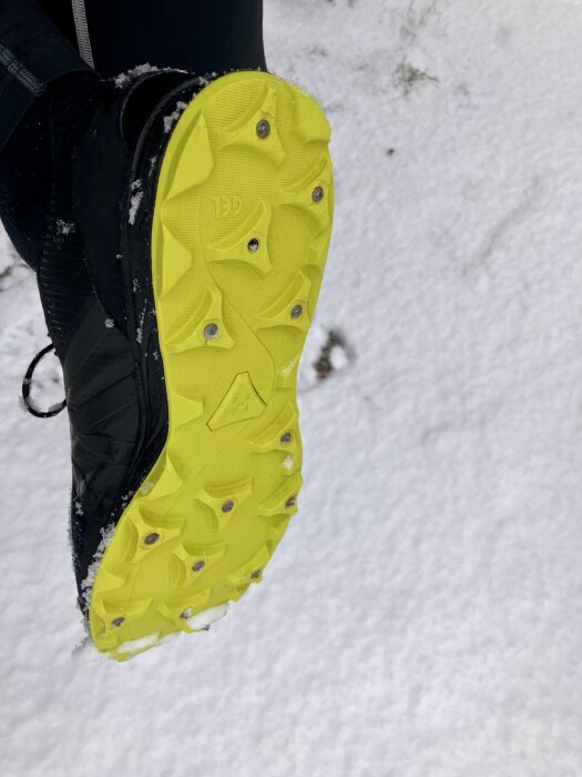 En närbild av en svart sko med gul sula utrustad med dubbar för halkskydd på snötäckt mark.