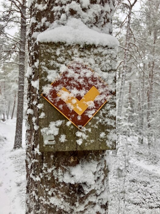 Snötäckt trädstam med en orange och gul markering på en skylt i en snöig skog.