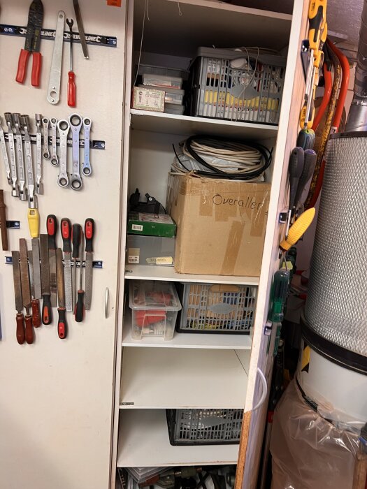 Verkstadsskåp med handverktyg på dörren, hyllor med plastkorgar och lådor, samt en kartong märkt "Overall". Dammsugare syns delvis till höger.