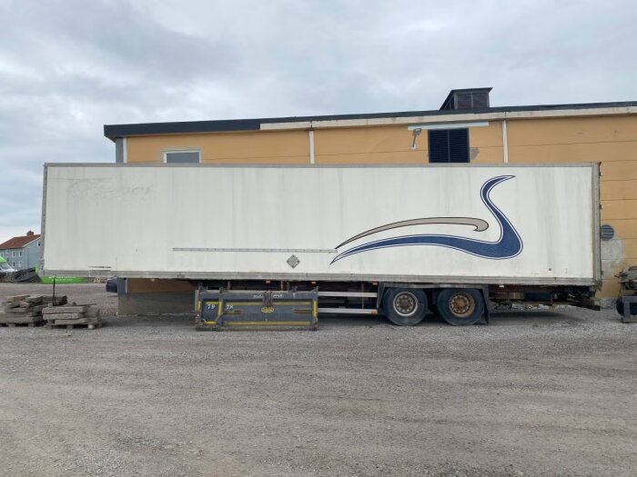 Vit släpvagn med blå dekoration parkerad framför ett gult hus, omgivet av grus och några staplade träpallar.