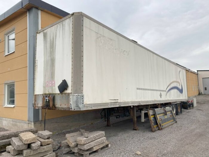 Vit trailer parkerad bredvid en byggnad, med betongblock framför och enkla dekorationer på sidan.