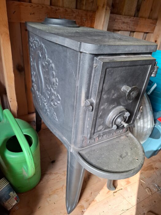 Gammal vedkamin med detaljerad design, placerad på trägolv bredvid en grön vattenkanna, i en träbyggnad.