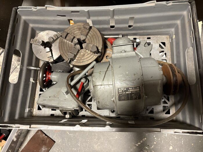 Äldre metallsvarvstillbehör och motor i en plastlåda, inklusive chuckar och ASEA-motor.