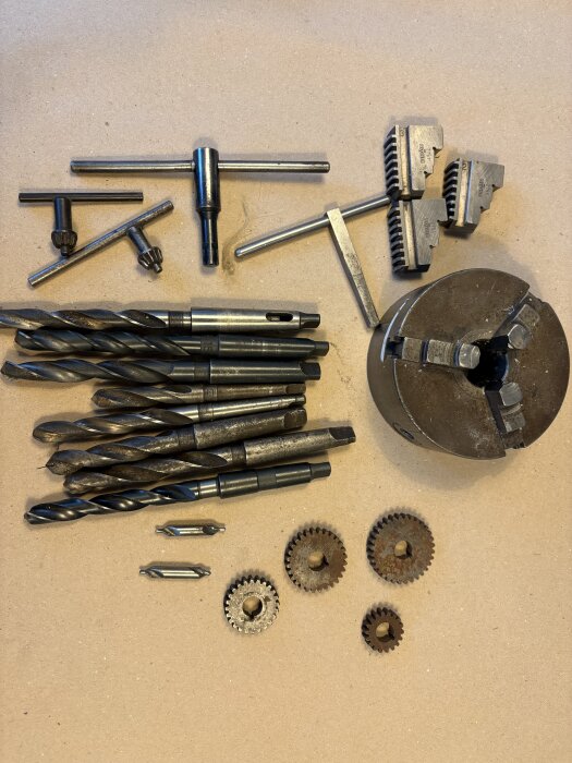 Metallsvarvstillbehör inklusive borrstål, chuckar, kugghjul och nycklar, utlagda på ett bord.