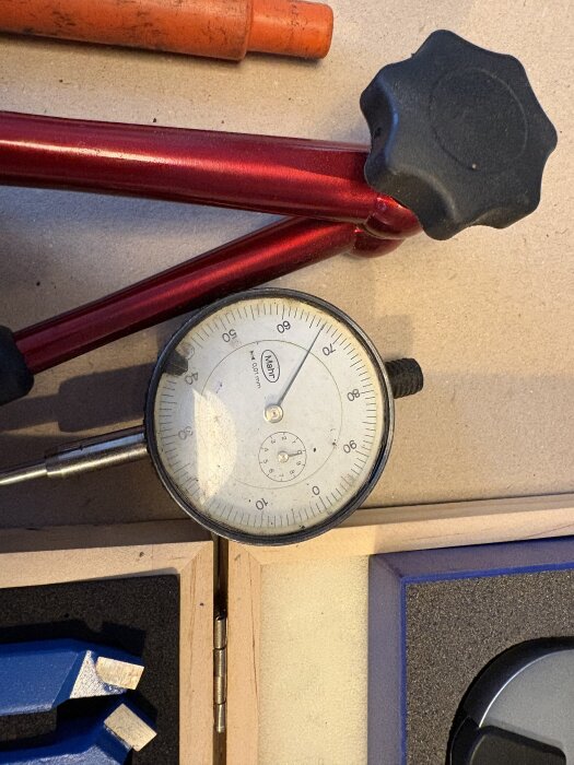 Mätklocka med stativ i röd metall och svart justerskruv, placerad på ett bord med andra verktyg.
