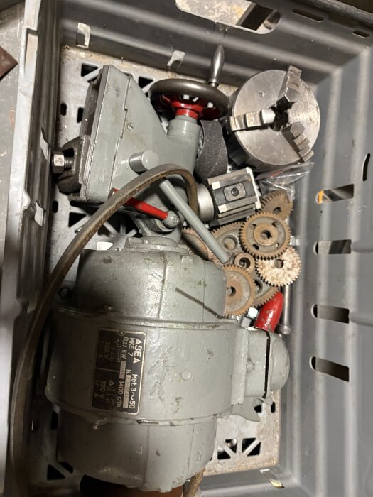 Äldre metallsvarvsdelar i en låda, inklusive motor, kugghjul, chuck och tillbehör för svarvstål.