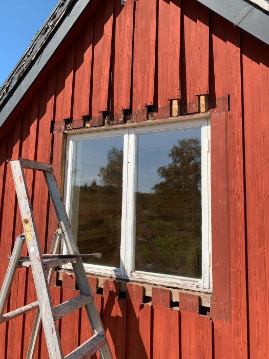 Byggnad med rött träfasad och nytt tvåluftsfönster. En del av fasaden är borttagen, samt en stege lutad mot väggen.