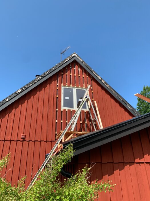 Röd träfasad av ett hus under renovering, stege lutad mot väggen, antenn på taket, tydlig blå himmel i bakgrunden.