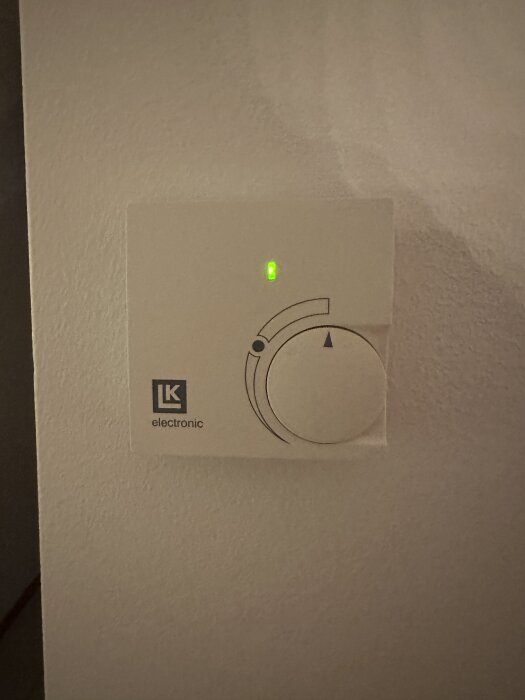 Rumstermostat med inställningsknapp och grön indikatorlampa monterad på vägg.