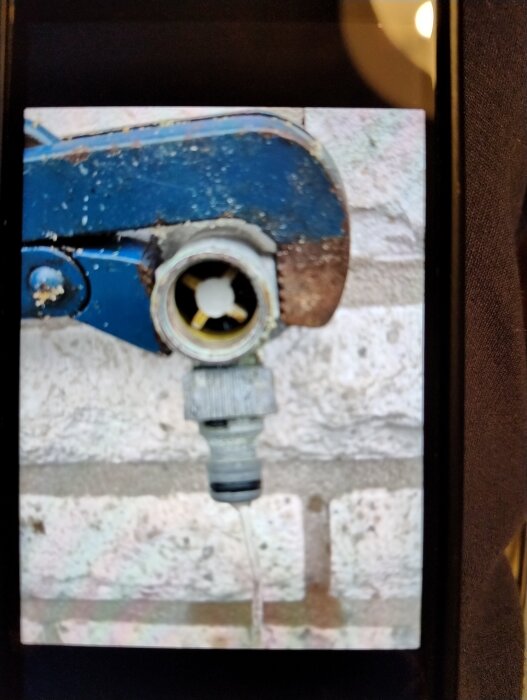 En blå och rostig vattenkran med en fastmonterad slangkoppling, fotad mot en vit tegelvägg.