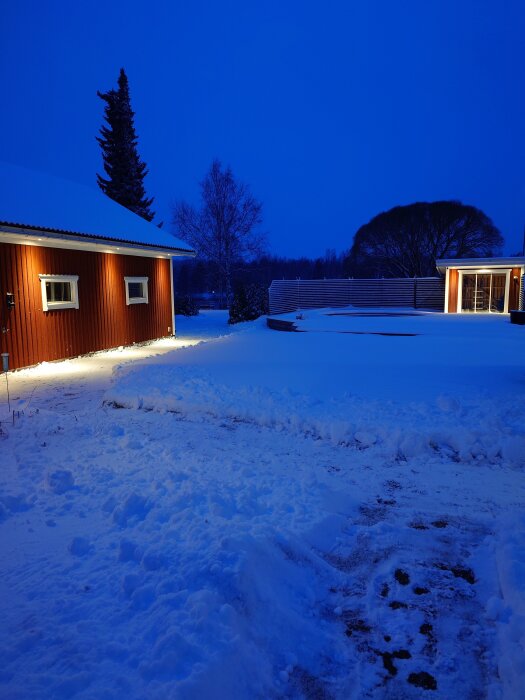 Snöigt landskap med belysta röda byggnader i skymningen, omgivna av snötÀckta trÀd och staket.