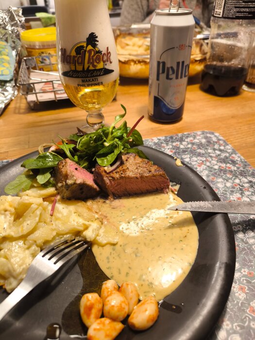 Oxfilé med potatisgratäng och bearnaisesås, sallad och en öl från Hard Rock Café i glas bredvid en burk Pelle-öl på ett bord.