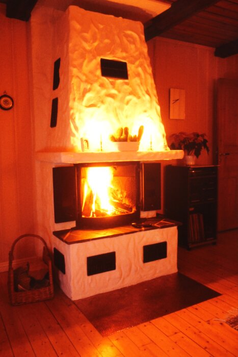 Öppen spis med eld i brinnande ved, luftutsläpp ovanför och runt kamin. Korg på golvet bredvid, med värme och eld reflekterande i rummet.